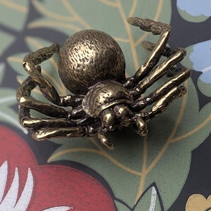 Könnte beinhalten: Ein bronzefarbenes, spinnenförmiges Dekorationsobjekt. Die Spinne hat einen strukturierten Körper und Beine mit detailliertem Design. Sie befindet sich vor einem Hintergrund aus floralen Mustern in Rot, Grün und Lila.