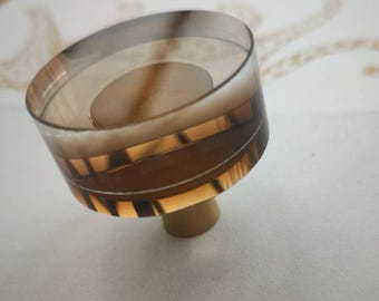 Könnte beinhalten: Ein dekorativer, bernsteinfarbener Glasknauf mit zylindrischer Form. Der Knauf hat horizontale Bänder in verschiedenen Braun- und Goldtönen. Der Sockel des Knaufs ist hellbeige.