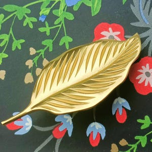 May include: A gold-coloured leaf-shaped brooch with detailed veining. The brooch is set against a dark background with a floral pattern in red, blue, and green.