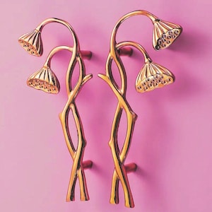 May include: A pair of gold-coloured cabinet pulls, each with a floral design. The pulls have a stem-like structure with a curved top and a flower-like head with detailed perforations. They are mounted on a pink background.