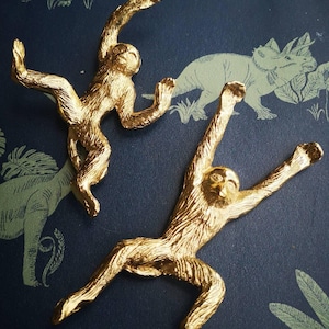 May include: Two gold-colored monkey figurines in dynamic poses. The monkeys are set against a dark blue background with a subtle pattern of dinosaurs and foliage. The figurines have detailed textures, capturing the essence of the animal.