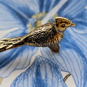 May include: A detailed, golden-colored bird figurine with textured feathers and a branch-like base. The bird is perched against a backdrop of blue and white floral patterns, creating a nature-inspired aesthetic.
