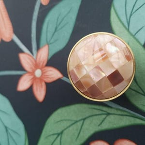 Puede incluir: Un pomo de armario redondo con una superficie similar a un mosaico de cuadrados marrones claros y rosas, engastado en un marco de metal dorado. El pomo está sobre un fondo oscuro con un estampado floral de flores color coral y hojas verdes.