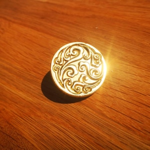 May include: A gold-colored, circular cabinet knob with an ornate, swirling design. The knob is set against a warm, wooden surface, with a bright light reflecting off the knob's surface.