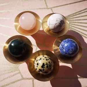 May include: Five gemstone spheres in brass bowls on a pink surface with gold lines. The spheres include rose quartz, white marble, green jasper, blue sodalite, and dalmatian jasper. The bowls are circular with a shallow depth.