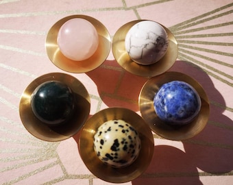 May include: Five gemstone spheres in brass bowls are arranged on a pink surface with gold lines. The spheres include rose quartz, white marble, green jasper, blue sodalite, and dalmatian jasper. The bowls are circular with a shallow depth.