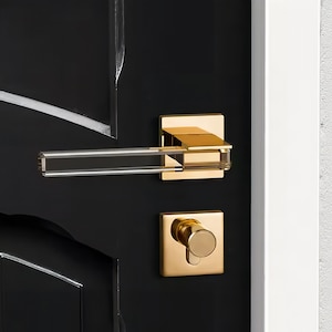 May include: Gold door handle with a clear glass lever and a square knob. The handle is mounted on a black door.