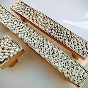 May include: A set of gold-toned cabinet hardware, including two rectangular pulls and a square knob. The hardware features a textured, pebble-like design within a rectangular frame, creating a luxurious aesthetic. Ideal for kitchen or bathroom cabinets.