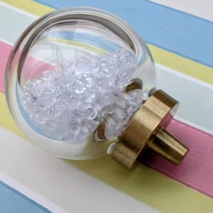 May include: A clear glass knob filled with small, clear, faceted crystals. The knob has a brass-colored base and stem. The background is a pastel striped pattern in pink, yellow, blue, and white.