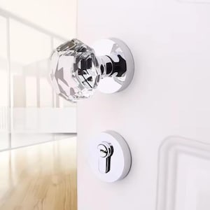 Puede incluir: Un pomo de puerta cromado con una placa frontal de cristal transparente. El pomo está montado en una puerta blanca. La puerta tiene un pestillo cromado a juego.