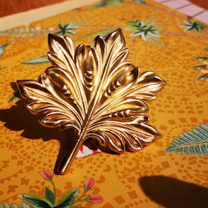 May include: A gold-coloured leaf-shaped brooch with detailed veining. The brooch has a stem and is set against a yellow floral patterned background. The brooch is likely made of metal and has a shiny finish.