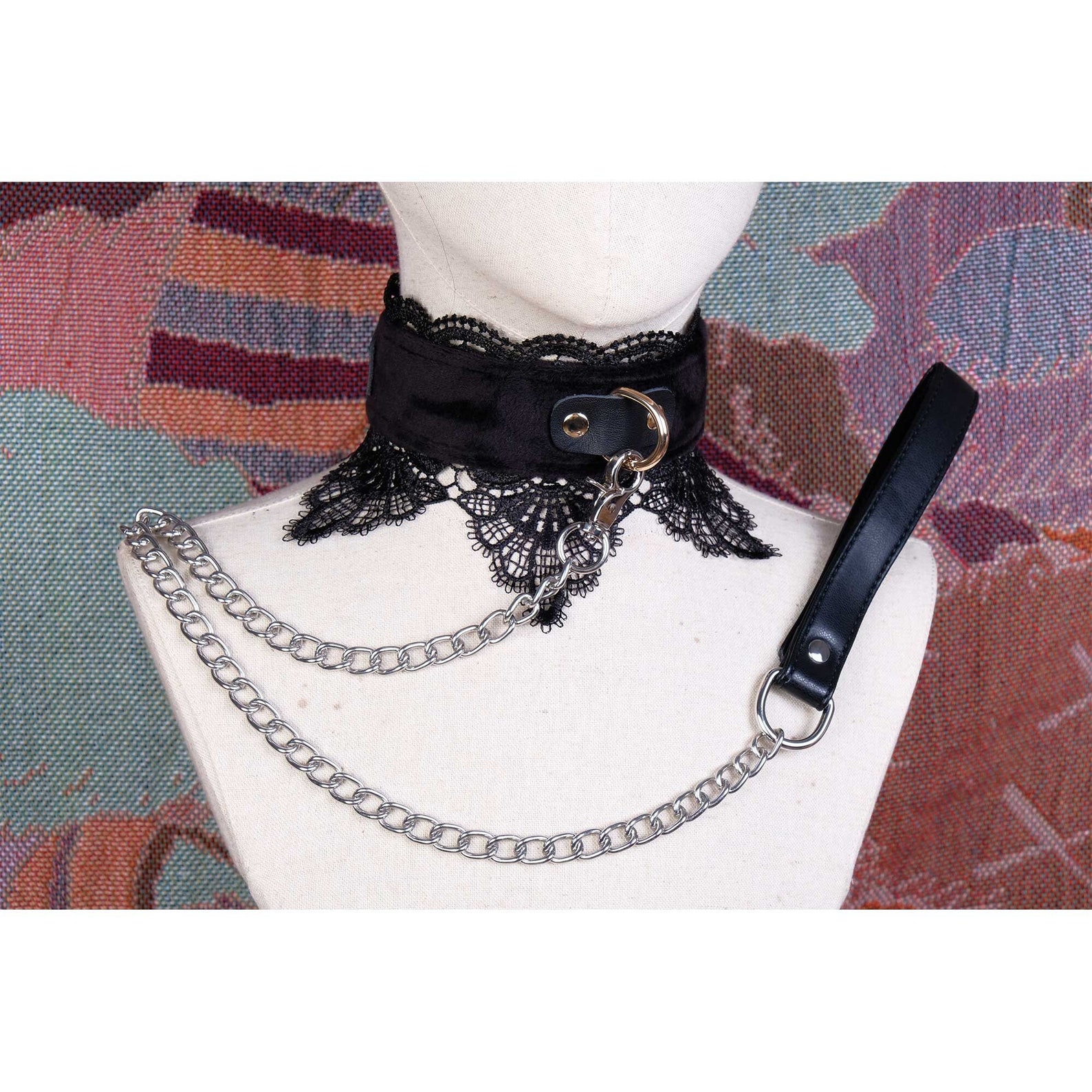 Black lace bdsm collar with soft faux fur lining and silver chain leash displayed on mannequin against colorful textile background.