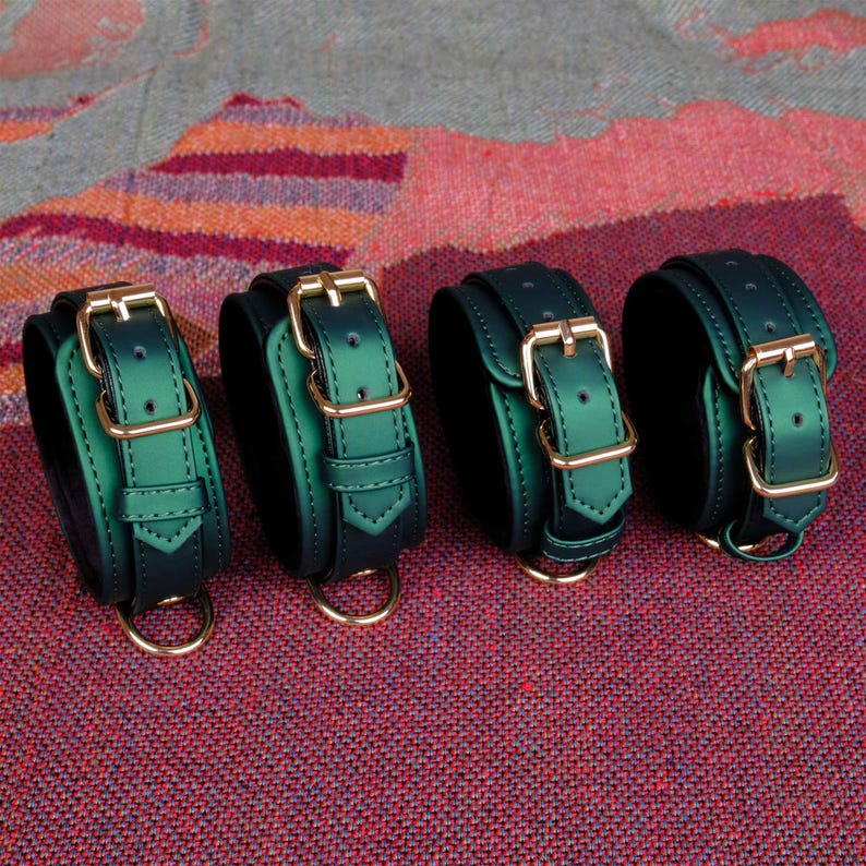 May include: Four green leather wrist cuffs with gold buckles and D-rings. The cuffs are arranged in a row on a red and orange patterned surface.