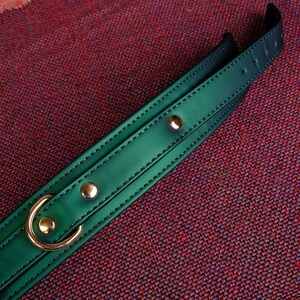 May include: A dark green leather collar with gold metal hardware. The collar has a stitched edge and two gold metal rivets.