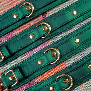 May include: Green leather straps with gold buckles and gold studs. The straps are arranged in a criss-cross pattern on a colorful fabric background.