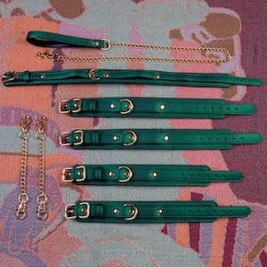 May include: Five green leather collars with gold hardware and two gold chains with clasps. The collars are arranged in a vertical stack on a patterned surface.