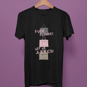 May include: Dark gray t-shirt with a graphic of three handbags in gray, pink, and beige. The text "FUCK FLOWERS, GET ME A BIRKIN" is printed in pink above and below the handbags. The shirt is hanging on a wooden hanger.