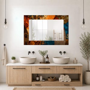 May include: A rectangular wall mirror featuring a decorative border with a patchwork design of orange, brown, and blue floral and geometric patterns. The mirror reflects a bright room with a window and a towel hanging on a wooden ladder rack.