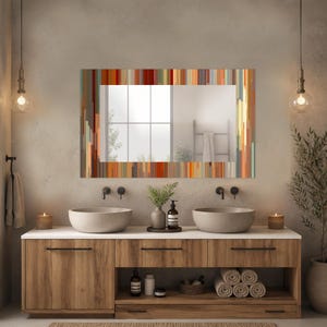 May include: A rectangular bathroom mirror with a colorful, vertical striped frame. The mirror hangs above a wooden vanity with two vessel sinks and black faucets. The room has a neutral color palette with warm lighting.