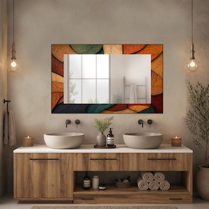 May include: A rectangular wall mirror featuring a decorative frame with a layered leaf pattern in shades of orange, brown, teal, and beige. The mirror is positioned above a wooden vanity cabinet with two round stone sinks and black faucets.