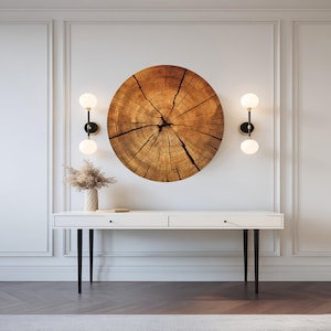 May include: A round wooden wall art piece with a detailed cross-section design hangs above a white console table. The table has black legs and two drawers. Two sconces with spherical lights flank the artwork.