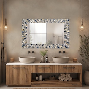 May include: A rectangular bathroom mirror with a mosaic tile frame in shades of blue, gray, and gold. The mirror is mounted above a wooden vanity with two vessel sinks and a white countertop. The room has a modern, spa-like aesthetic.