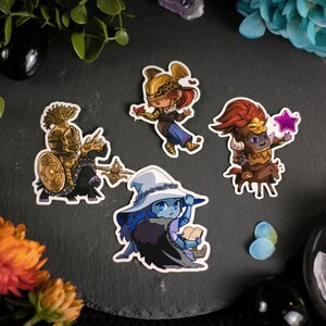 Può includere: Una collezione di cinque adesivi di personaggi a tema fantasy. Gli adesivi raffigurano varie figure in armature e abiti dettagliati, con colori vivaci e tratti da cartone animato. Gli adesivi sono disposti su una superficie scura e strutturata, circondati da elementi floreali e decorativi.