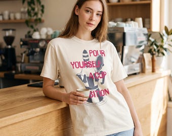 May include: A cream-colored short-sleeve t-shirt featuring a graphic of a moka pot and the text "POUR YOURSELF A CUP OF AMBITION" in pink lettering. The shirt has a crew neckline and a relaxed fit, worn with light blue denim jeans.