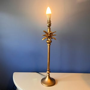 May include: A tall, gold-colored metal table lamp featuring a slender, cylindrical stem and a circular base. The lamp includes a decorative pineapple-shaped element near the top, below the light bulb socket, which holds a single lit candle-style bulb.