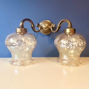 May include: Vintage double wall light with clear glass shades etched with a floral design. The sconce has a brass-toned metal frame and mounting plate. The shades have a scalloped edge.