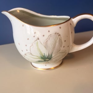 May include: White ceramic creamer with a gold rim and handle. The creamer features a floral design with a gold polka dot pattern. The interior has a light green colour.