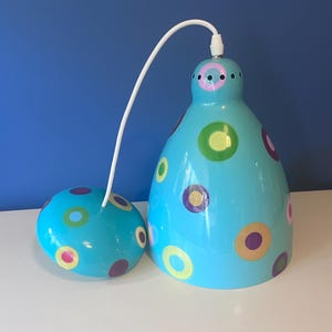 May include: A turquoise pendant light fixture with a matching ceiling mount. The light has a rounded shape with colorful polka dots in yellow, green, and purple. A white cord connects the two pieces.