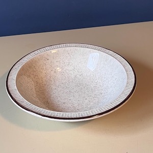 May include: A speckled, off-white ceramic bowl with a dark brown rim and a textured, ridged border. The bowl is round and sits on a light-coloured surface, with a dark blue background visible.