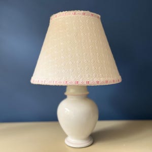 May include: A white ceramic table lamp with a cream lampshade. The lampshade features a lace pattern and pink ribbon trim. The lamp base is rounded. The lamp sits on a cream surface against a blue backdrop. A decorative lighting fixture.