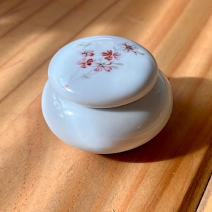 May include: A small, round white porcelain trinket box with a smooth, glossy finish. The lid features a delicate hand-painted floral motif in shades of pink and green, depicting a cluster of small blossoms with thin, trailing stems and leaves.