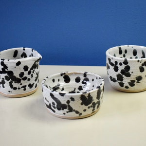 May include: A ceramic set consisting of a creamer jug with a handle and spout, a small round bowl, and a mug with a handle. Each piece features a white base with an irregular black speckled pattern and a smooth, glossy finish throughout.