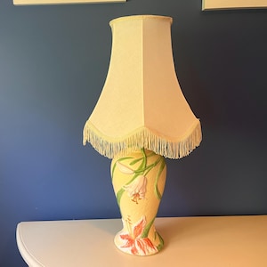 May include: A table lamp with a cream-coloured fabric shade featuring a fringed bottom edge. The ceramic base is pale yellow with a textured surface, decorated with raised, hand-painted lily flowers in shades of white, pink, and green foliage.