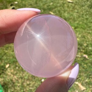May include: A translucent, light pink crystal ball, held between fingers against a backdrop of green grass. The sphere has a smooth surface with internal light reflections, creating a starburst effect. The crystal is a soft, pastel pink.