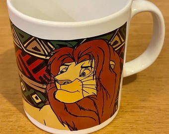 Op de afbeelding: Witte keramische mok met een kleurrijk beeld van Simba uit Disney's The Lion King. De afbeelding toont Simbas gezicht en manen, met een groene, rode en gele achtergrond.