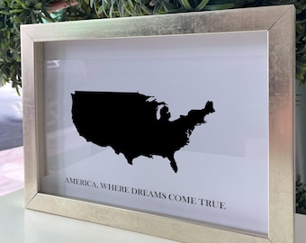 May include: A rectangular silver-toned frame containing a white print with a solid black silhouette of the United States map. Below the map, the text reads: AMERICA, WHERE DREAMS COME TRUE. The frame has a metallic finish and a clean, simple design.
