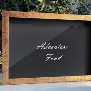 Könnte beinhalten: Ein rechteckiger Schattenfugenrahmen mit einem abgenutzten Goldfinish und einer schwarzen Innenrückwand. Die Glasfront zeigt den weißen Schriftzug Adventure Fund in Schreibschrift, der mittig im Rahmen für eine klare Sicht platziert ist.