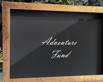 May include: A rectangular shadow box frame with a distressed gold finish and a black interior backing. The glass front features the white text Adventure Fund printed in a cursive script, centered within the frame for a clear and readable display.