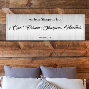 May include: A white wooden sign with black text that reads "As Iron Sharpens Iron One Person Sharpens Another Proverbs 27:17" hanging on a rustic wood wall above a bed with gray and white bedding.