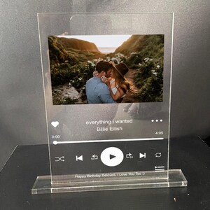 May include: A clear acrylic music player stand with a photo of a couple kissing in a forest. The music player screen displays the song "everything i wanted" by Billie Eilish. The text "Happy Birthday Beloved, I Love You Too" is printed on the base of the stand.