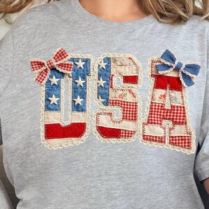 May include: A heather gray short-sleeve t-shirt featuring the letters USA in a patchwork style. The U is blue with white stars, the S is a floral pattern, and the A is red and white plaid. Two small fabric bows are attached to the top letters.