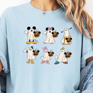 May include: A light blue t-shirt with a graphic of Mickey Mouse, Minnie Mouse, Donald Duck, Goofy, and Pluto dressed as ghosts holding jack-o-lanterns. The characters are in a row, with Mickey Mouse on the left and Pluto on the right.