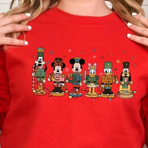 May include: A red long-sleeve sweatshirt featuring a colorful graphic of Disney characters dressed as nutcrackers. The characters include Mickey Mouse, Minnie Mouse, Donald Duck, and Goofy. The design is festive and holiday-themed.