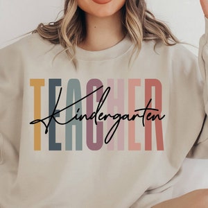 K&ouml;nnte beinhalten: Ein hellbeiger Pullover mit dem Text "TEACHER kindergarten" in bunten vertikalen Streifen. Der Text ist in einer modernen, handschriftlichen Schrift.