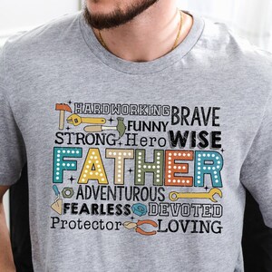 May include: Gray t-shirt with a graphic design featuring the word "Father" surrounded by words describing positive qualities, such as "hardworking", "brave", "funny", "strong", "hero", "wise", "adventurous", "fearless", "devoted", "protector", and "loving".
