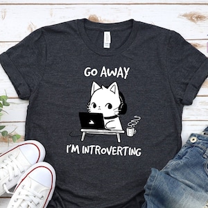 May include: Dark heather grey t-shirt with a white cat wearing headphones and using a laptop. The text on the shirt reads "Go Away I'm Introverting".